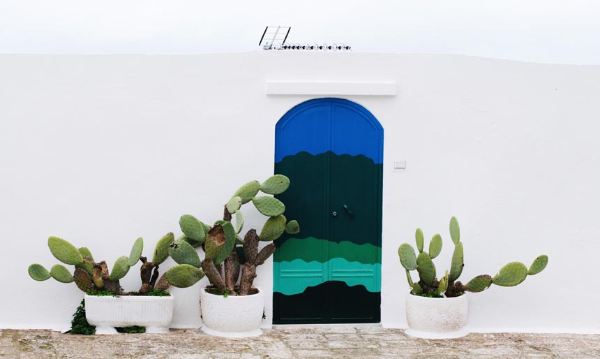 An entrance door painted in various blue toned hues against an off white facade and cacti on either side of it.