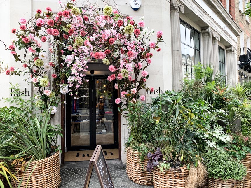 An entrance to a store with huge basket planters to either side of it and a canopy of roses over it.