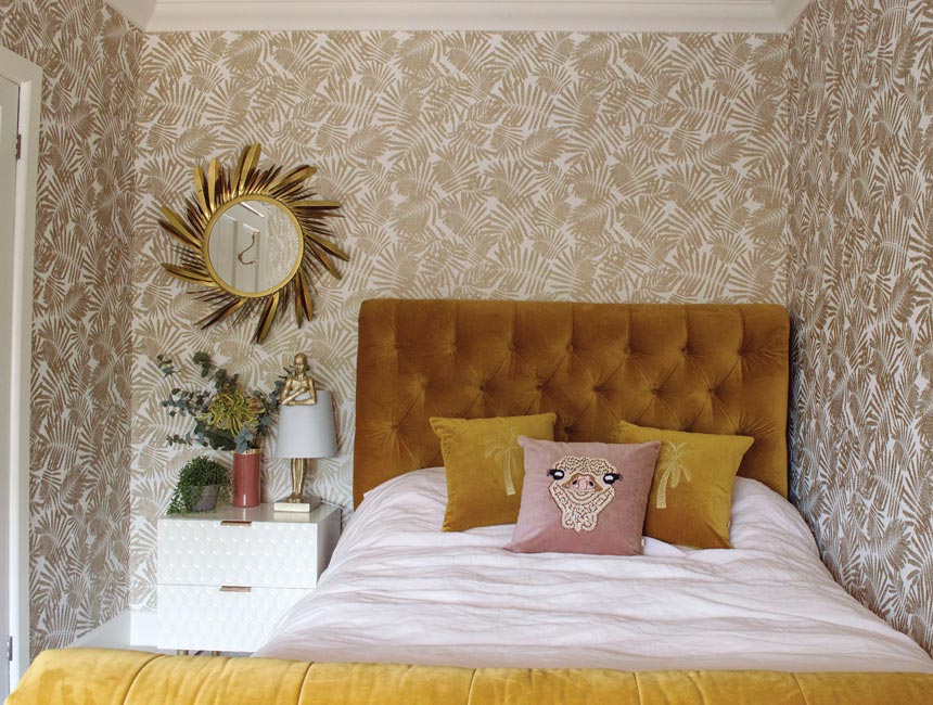 A bedroom with an gold leaf pattern wallpaper, a mustard hue bed and decorative pillows on top of the white bedding. Image via Audenza.