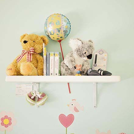 A white shelf against a light green wall with decor for babies (little books and cuddle toys) as part of a nursery