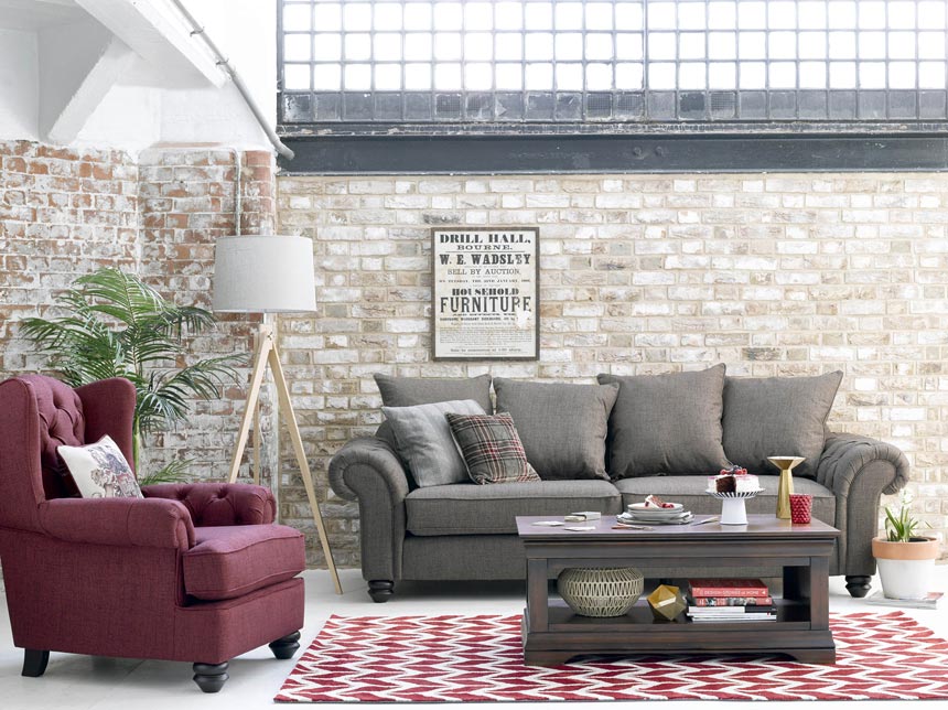 An interior with a gray sofa, a red wine armchair, a floor lamp in between, an area rug with a red wine chevron pattern and a brick accent wall. Image via Oakfurnitureland.