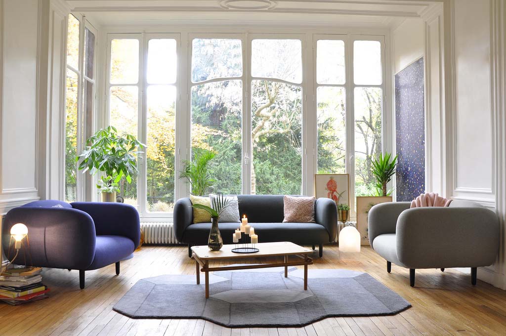 A very bright living room with a gray three seater sofa and two armchairs with a coffee table in the middle atop a irregular shaped rug. The background is a series of glass doors from floor to a very high ceiling. Image by ENO_Studio.