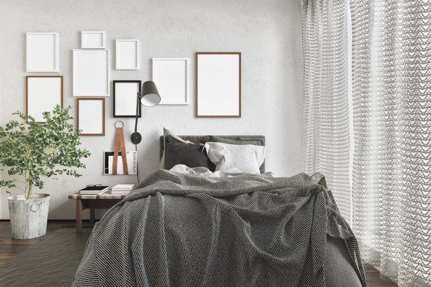 messy modern bedroom with an unmade bed and a series of blank picture frames on the wall lit by daylight from a large window with curtains