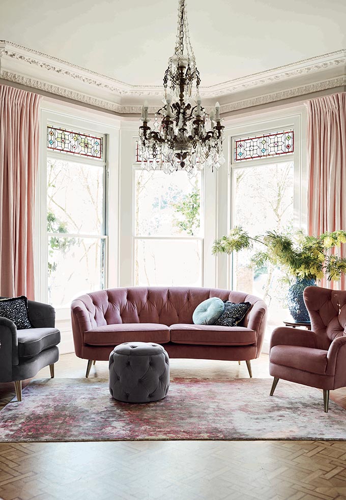 A pink curvy sofa with airy legs in between two other armchairs looking stylish and poised in this living room with a very high ceiling. Image by DFS Furniture.