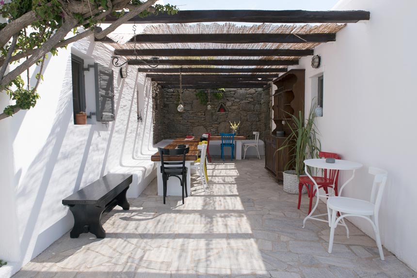 An outdoor dining setup sheltered by the sun via a pergola. In the background there is a masonry accent wall, pops of bright colors and a mix of different woods make up the rest of the decor in this Mykonian house.