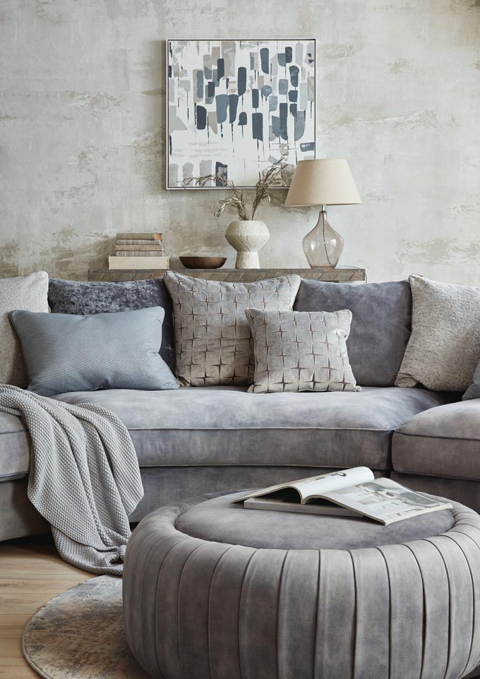 A beautiful curvy sofa, the Palm by Sofology, looking lushious with a whole lot of decorative pillows, a pouf and console table behind it. Image via Sofology.