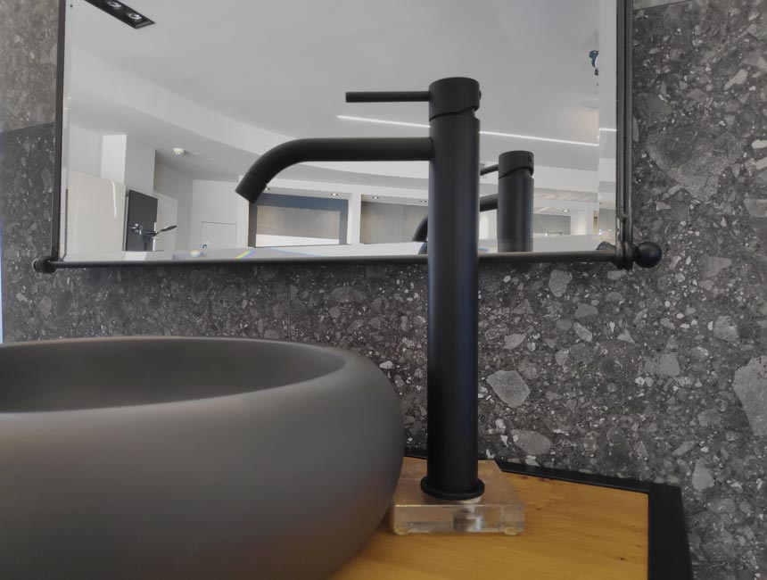 Detail of a wooden bathroom vanity with a black faucet, steel frame mirror and terrazzo backsplash.