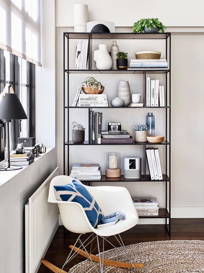 A contemporary styled vignette with a rack of bookshelves next to a window and an armchair. Image by John Lewis.