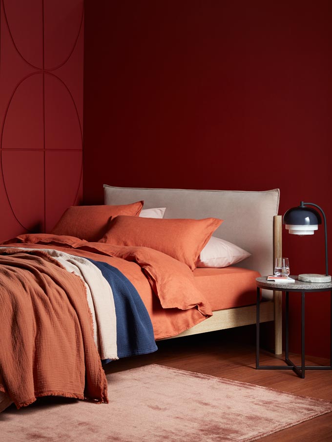 A deep saturated red bedroom corner with terracotta hue bedding and an off white bed looks impressive next to a terrazzo side table. Image by John Lewis.