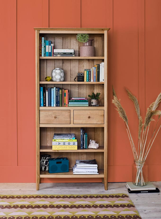 A solid pine bookcase that includes two drawers and decor besides book, against an terracotta hue accent wall. Image by Oakfurnitureland.