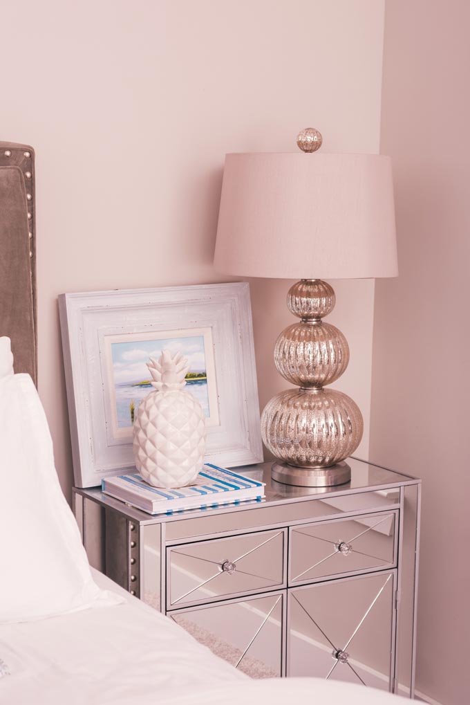 One of the six interior decorating fail proof tips calls for adding a rich paint color. A powder pink vignette by a bed with a mirrored nightstand, stylish decor and table lamp.