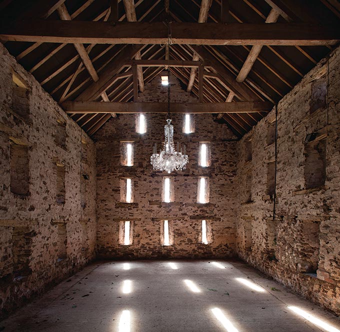 A gorgeous chandelier hanging from a timber truss sloped roof inside an old, empty stone building. Image by Fritz fryer.