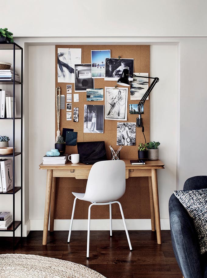 A home office vignette with a white desk chair, pinboard, wooden desk and black desk lamp. Image by John Lewis.