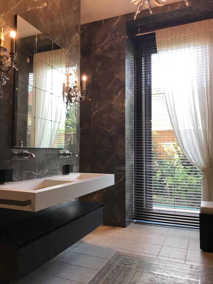 A luxurious bathroom with dark veined marble walls, a white sheer curtain and Venetian blinds.