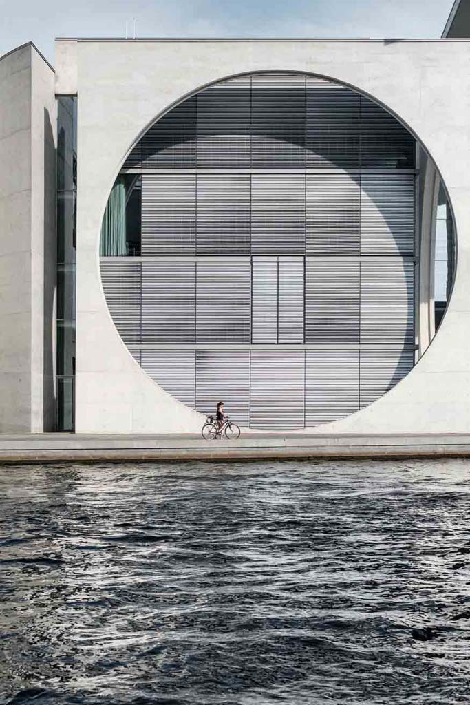 The exterior facade of a building with a tall concrete wall and a huge hole in it with large windows and Venetian blinds