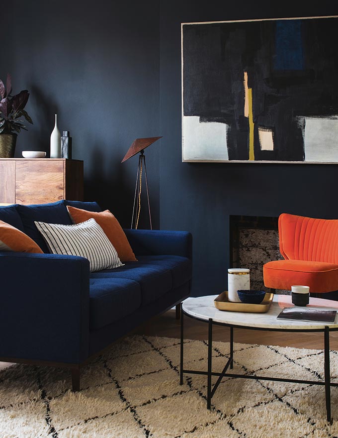 A moody blue living space with a large artwork, a blue sofa an orange armchair and three pillows, following the rule of 3's. That's one of the fail-proof interior decorating tips. Image: Swoon Editions.