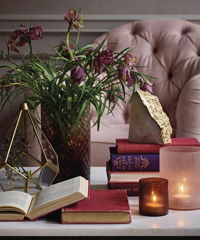 A close up of a white marble coffee table packed with tealights, books and a vase with flowers. In the background a blush pink armchair. Image by Marks & Spencer.