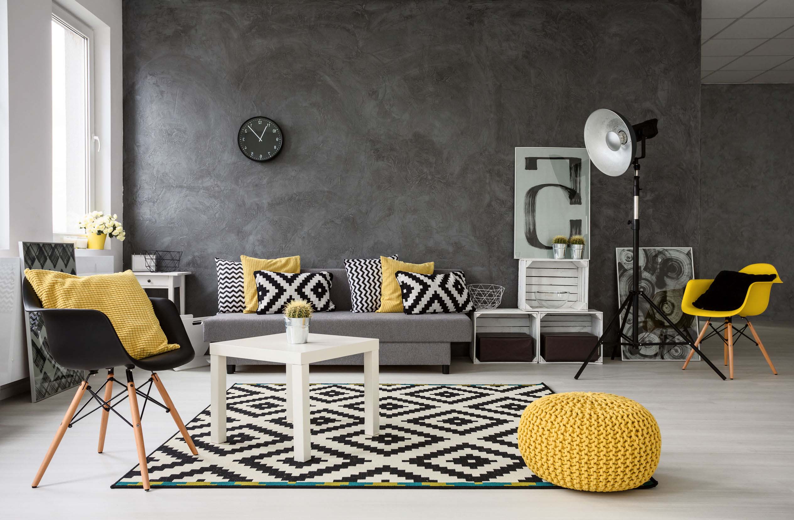 spacious, grey living room with sofa, chairs, standing lamp, small coffee-table, decorations in yellow, black and white. Behind the sofa there is a dark gray microcement accent wall