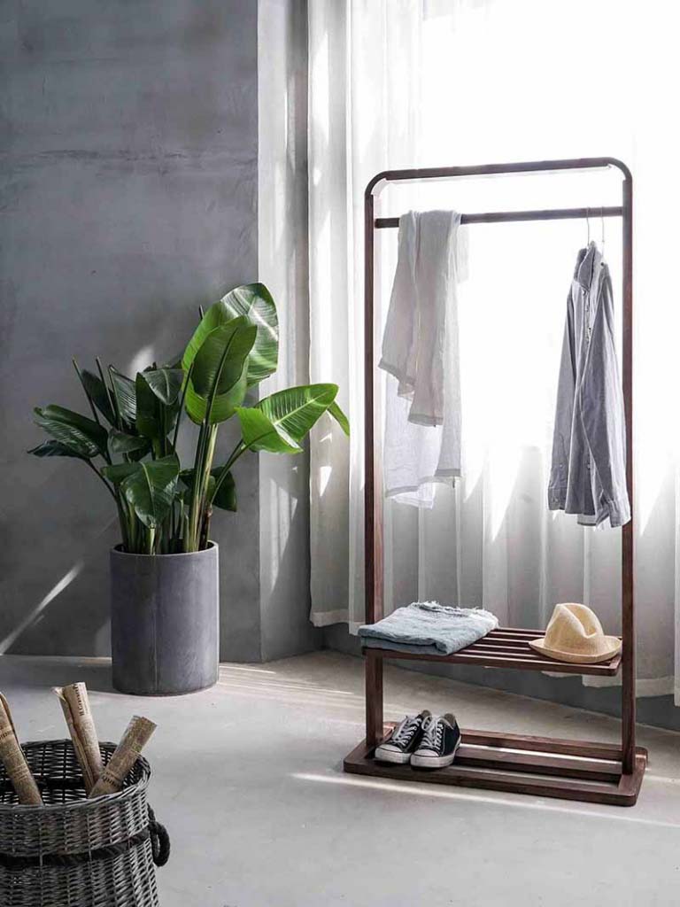 View of a room with a clothing rack in front of a window with white sheer curtain and a round tall concrete planter pot against a gray microcement wall