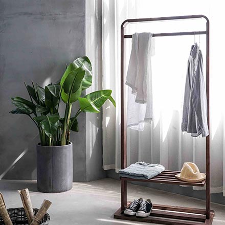 View of a room with a clothing rack in front of a window with white sheer curtain and a round tall concrete planter pot against a gray microcement wall