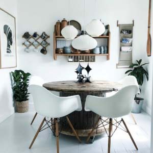 A white Scandinavian dining room filled vintage finds. There is a white three lamp pendant light hanging over a custom made rustic round table. There's also an open shelving unit on the wall with pots and mugs.