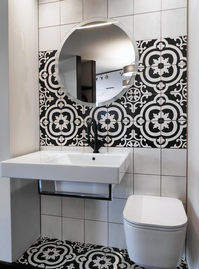 A contemporary bathroom with a white wall mount wash basin and toilet, a round mirror, white wall tiling and a decorative black pattern accent wall tiling zone and flooring to create that pop effect.