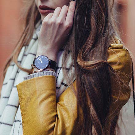 a partial view of a young brunette woman wearing a mustard colored leather jacket, with a white scarf around her neck and a black wrist watch