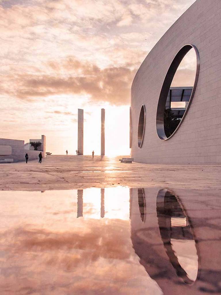 A modern building on the right against a pink cloudy sky in the background and a reflection of the sky on a paddle of water on the foreground. Everything has a pink hue