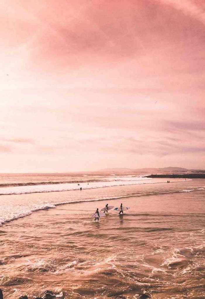 A pink sky at sunset hours by a beach with surfers where everything looks pink