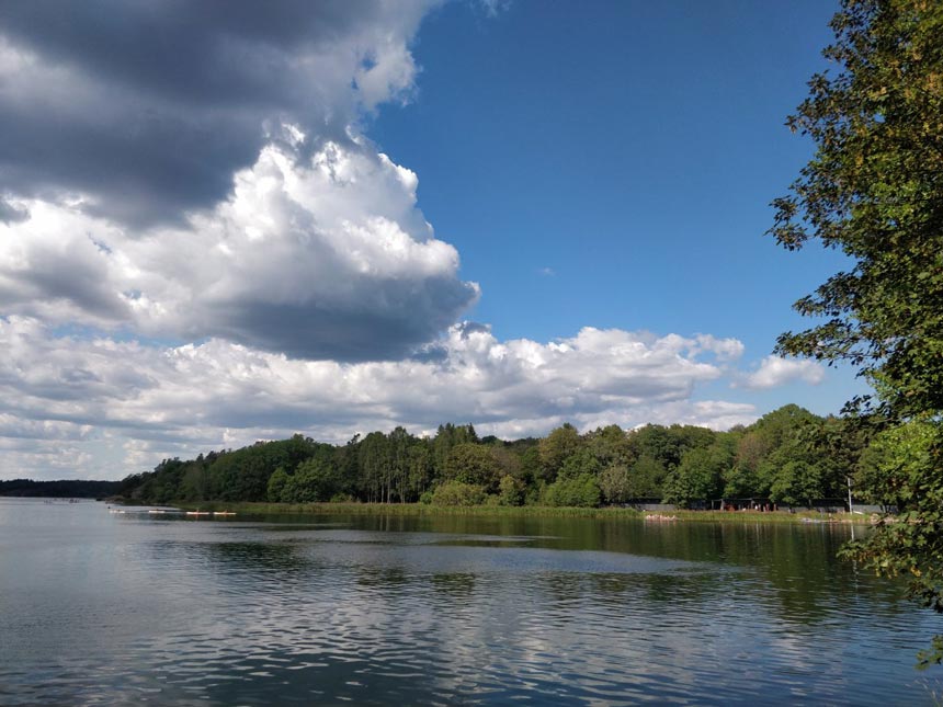 View of a lake with greenery on the one bank and some clouds over the blue sky. 