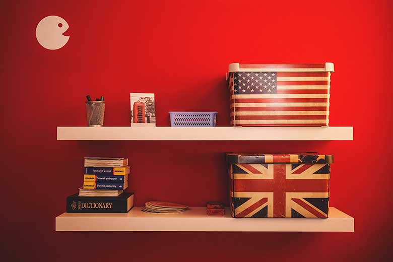 A red wall as a backdrop for two floating shelves with boxes, books and stationary on top of them. The two boxes have the American flag and the British flag on them.