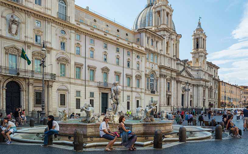 Palazzo Pamphili in Piazza Navona in Rome with a fountain in front of it and lots of passerbys