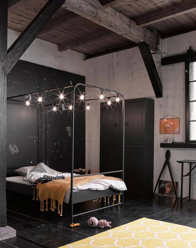 An industrial space with exposed dark wood beams has been styled as an industrial bedroom that has a canopy of Edison bulb lights. Image by Cuckooland.