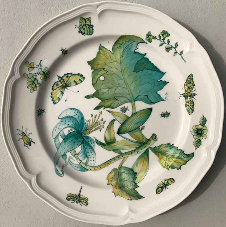 A dinner plate with a blue flower and lots of complimentary leaves and green bugs drawn on it