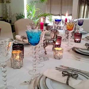 A beautifully styled tabletop for a New Year's Eve dinner party with lots of colored wine glasses, white porcelain plates with silver trims, silver cutlery all on a white tablecloth.