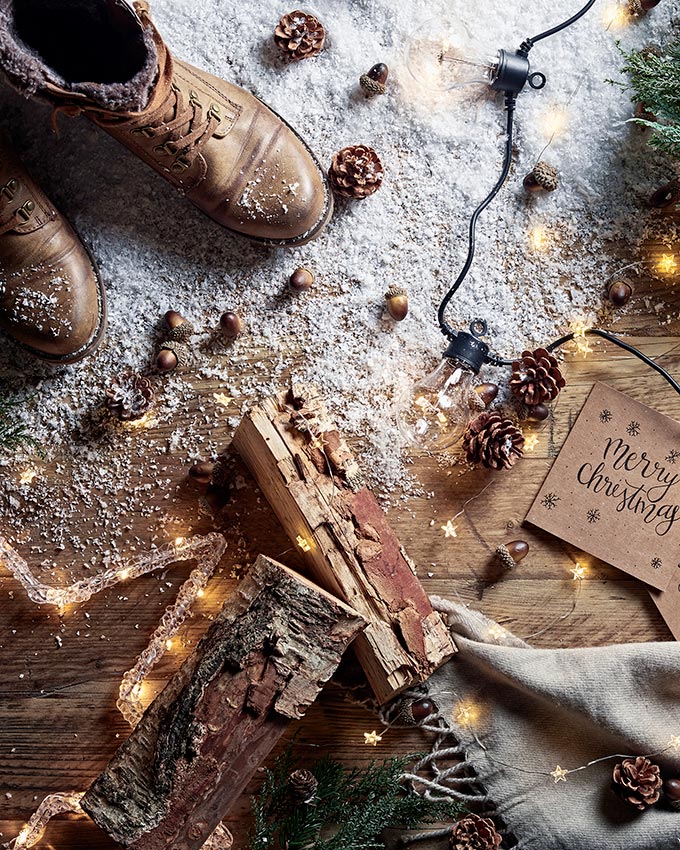 A flatlay with a pair of brown boots, timber logs, nuts and a string of lights on a wooden floor with sprinkles of snow. Image shot by Oliver Perrott for Lights4Fun.co.uk.