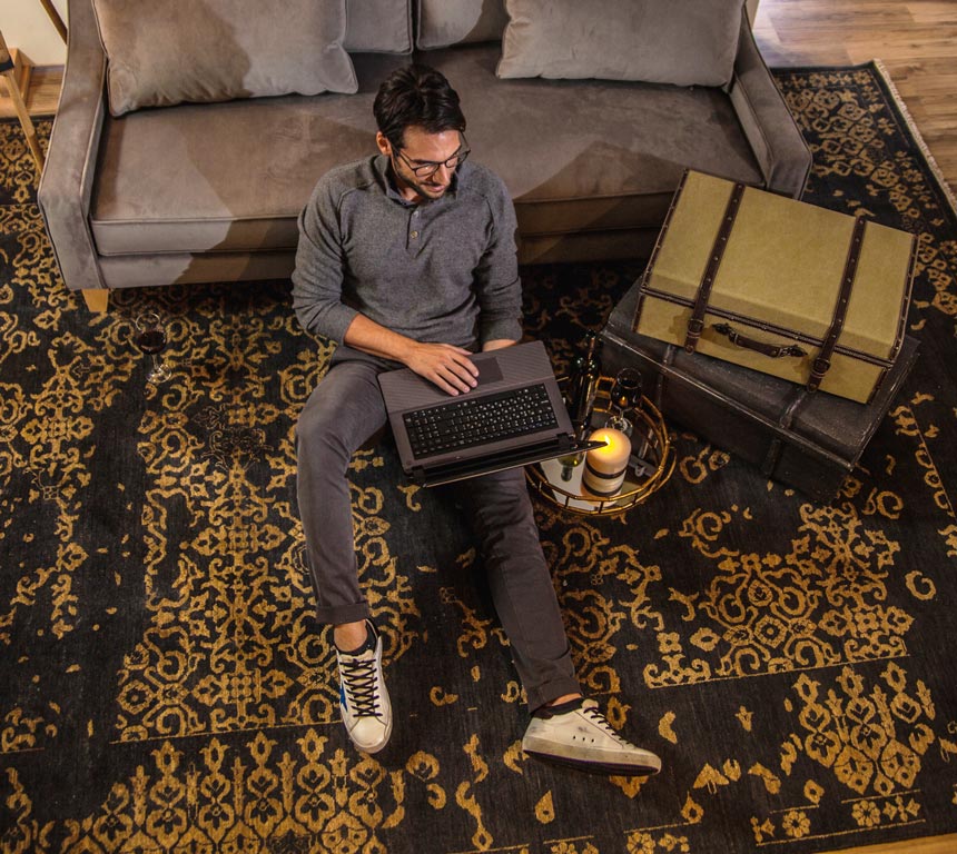 Anastasio Kyriakopoulos working on his laptop whilst sitting on a black area rug with a golden broken pattern, as part of a concept shoot.