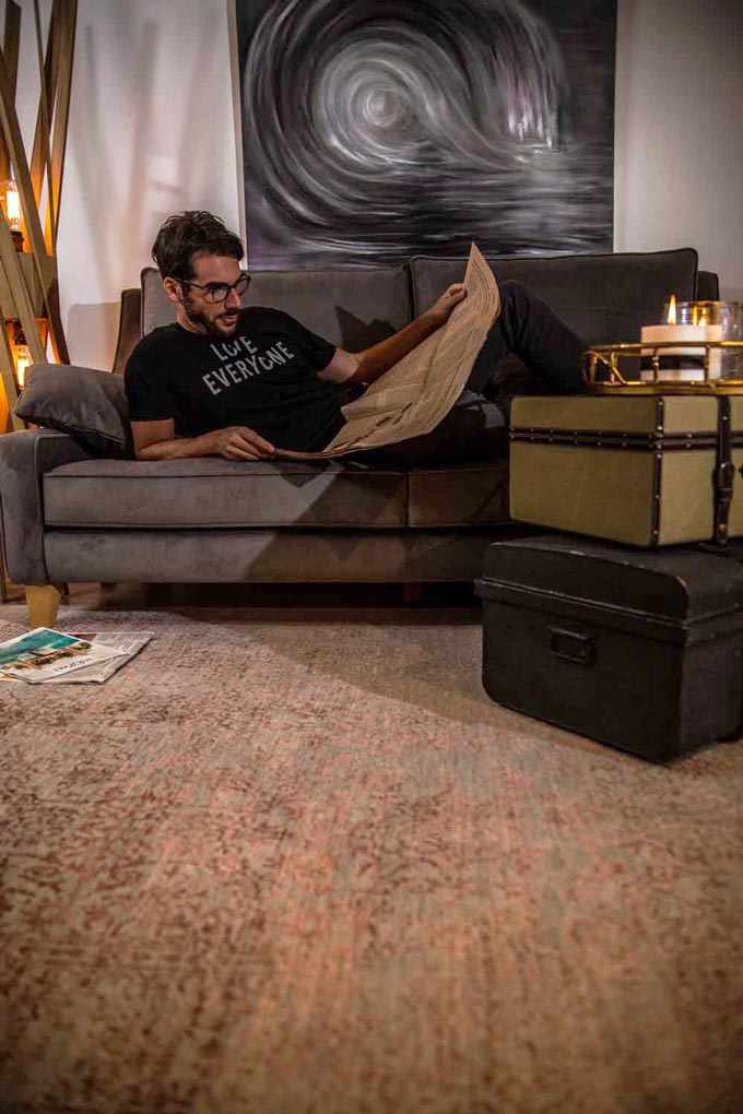 A male model lying on a velvet gray sofa reading a newspaper. Old suitcases are stacked up on the right with candles burning atop.