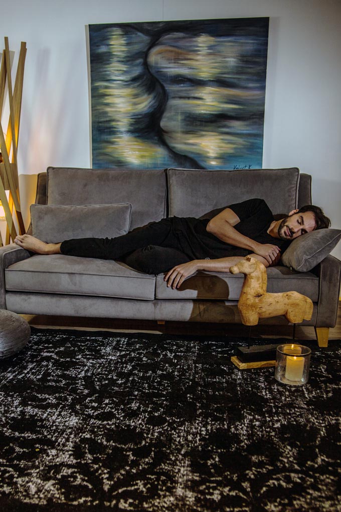 A man napping on a grey sofa. To the left there's a floor lamp. To the right front corner of the sofa, there's a wooden horse sculpture and a burning candle. A vintage black area rug sets the moody ambiance. On top of the velvet sofa, there's a large artwork