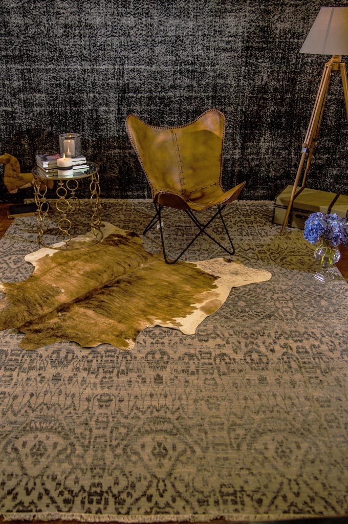 A brownish pattern area rug with a designer tanned leather armchair against a black accent wall with a brassy round side table and decor and a floor lamp on the either side. Setting from a concept shoot on masculine interiors.