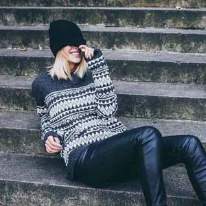 A blonde young woman with a hat on covering her eyes, is smiling while sitting on some concrete steps wearing a pattern sweater and black leather pants