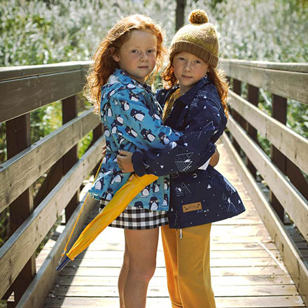 Two twin sisters looking pretty in raincoats embracing each other on a narrow timber footbridge outdoors.