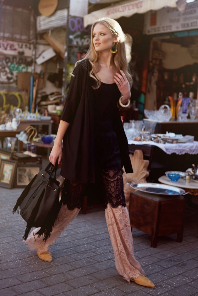 A beautiful young model wearing a black top with a pair of wide leg pants with lace, whose carrying a black leather bag is walking through a flea market looking gorgeous. All creations of RIEN by Penny Vomva.