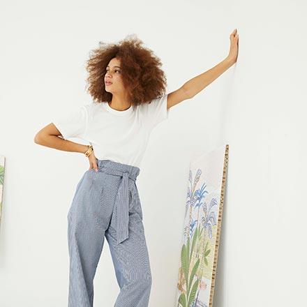 A woman with a white tee and blue high waisted pants leaning against a white wall. Image by Oliver Bonas.