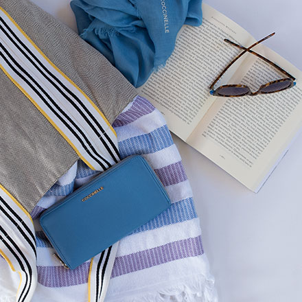 A Coccinelle wallet and peach scarf among a beach towel, sunglasses, open book and fabric beach bag. All casually laid flat. Image by Antonis Drakakis.