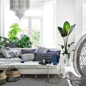 A white sitting room with a bohemian flair to it because of the white hanging Moroccan lantern, the seagrass baskets in front of the white sofa, the blue patterned throw pillows, the plants and the peackock fan white armchair. Image by Monsoon.