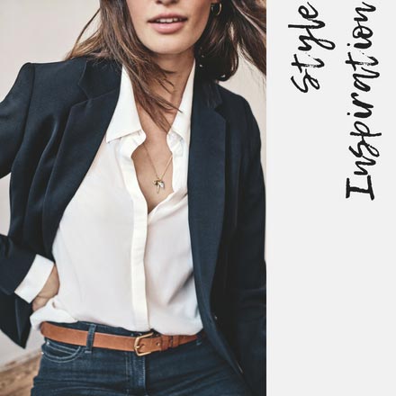 A close up of a chic woman dressed with an off white shirt, black tailored blazer jacket, and dark blue pants with a tanned thin leather belt around the waist. Image by PureCollection.
