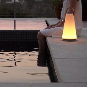A woman sitting by the edge of a pool with her feet inside the water and a portable light besides her after sunset hours creating a soothing ambiance. Image by Nest.co.uk.