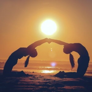 two girls reaching out their hand to hold each other while arching their backs on a beach with a sun about to set.