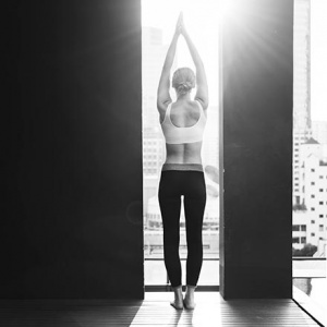 A black and white image of a woman in a sporty outfit with arms raised above her head looking out a narrow window that is from floor to ceiling and gazing at the cityscape.